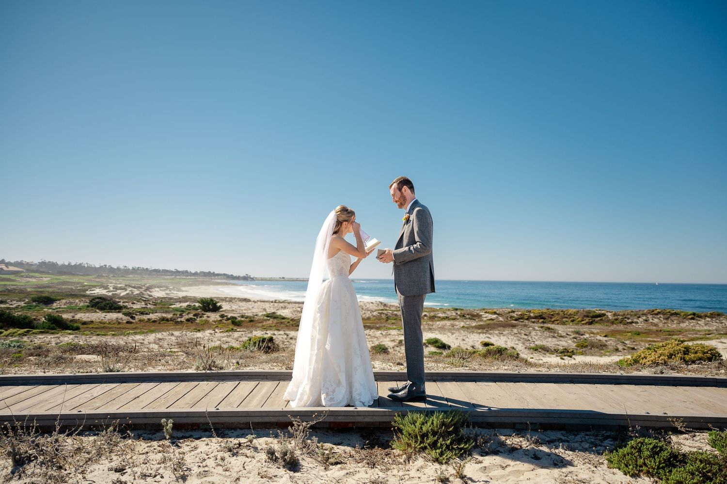 Bride and groom walk hand-in-hand on a sunny beach, bride in white gown with veil, groom in gray suit.