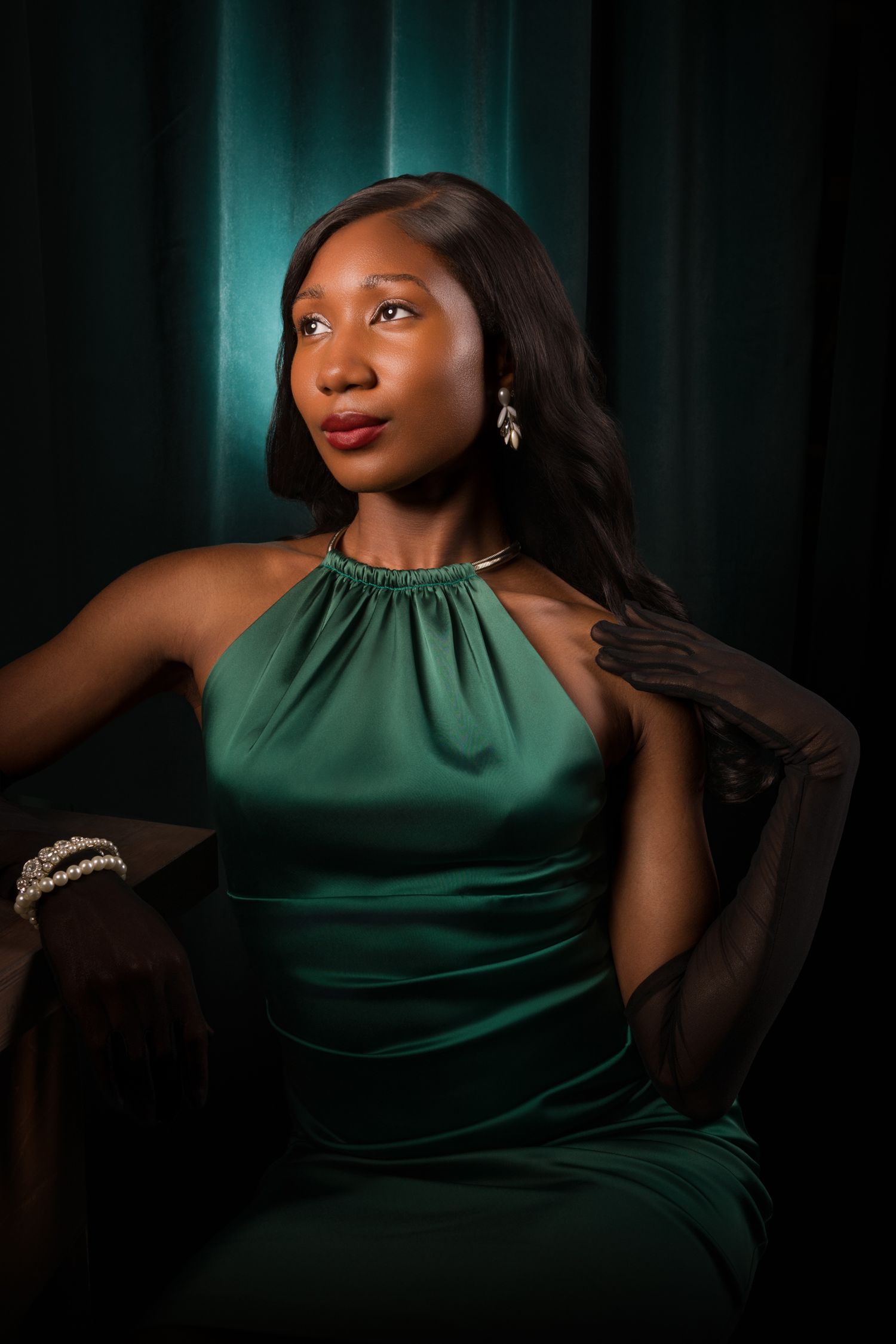 Elegant Black woman in emerald green satin halter gown with drop earrings and gloves, posed against teal curtain backdrop.