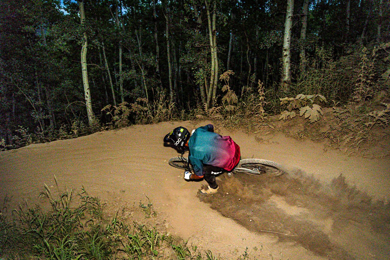 Mountain biker carving a fast berm turn on a dirt trail through a forest, leaning low with colorful gear.
