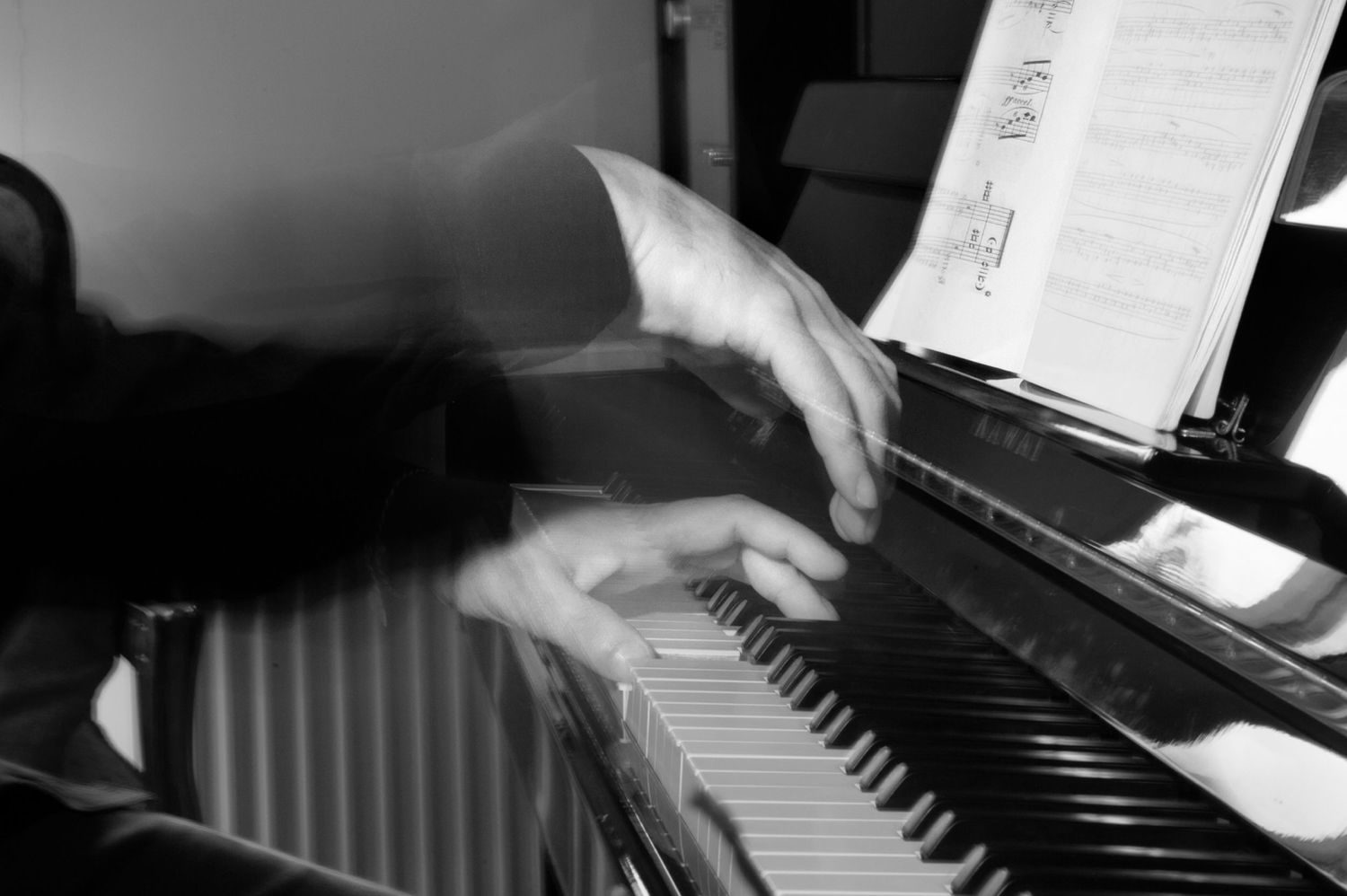 Blurred black and white image of hands playing a piano with sheet music, capturing motion and energy. The mood is artistic and dynamic
