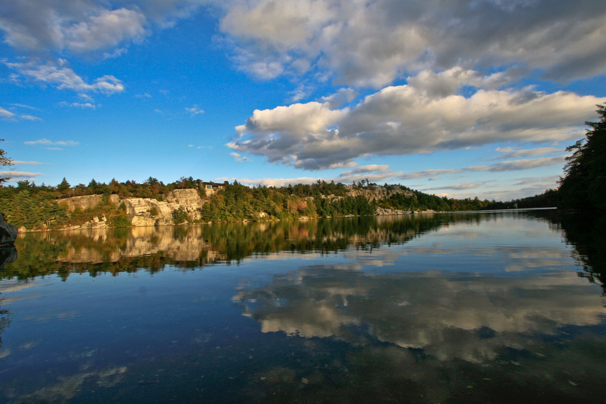 Calm lake reflecting blue sky and white clouds, surrounded by rocky cliffs and lush green trees in a serene landscape.