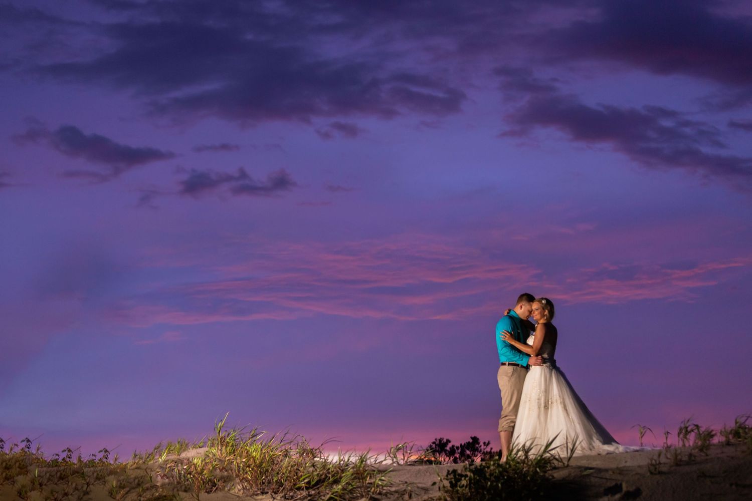 Couple embracing at sunset with vibrant purple and pink sky backdrop in a romantic outdoor wedding photo.