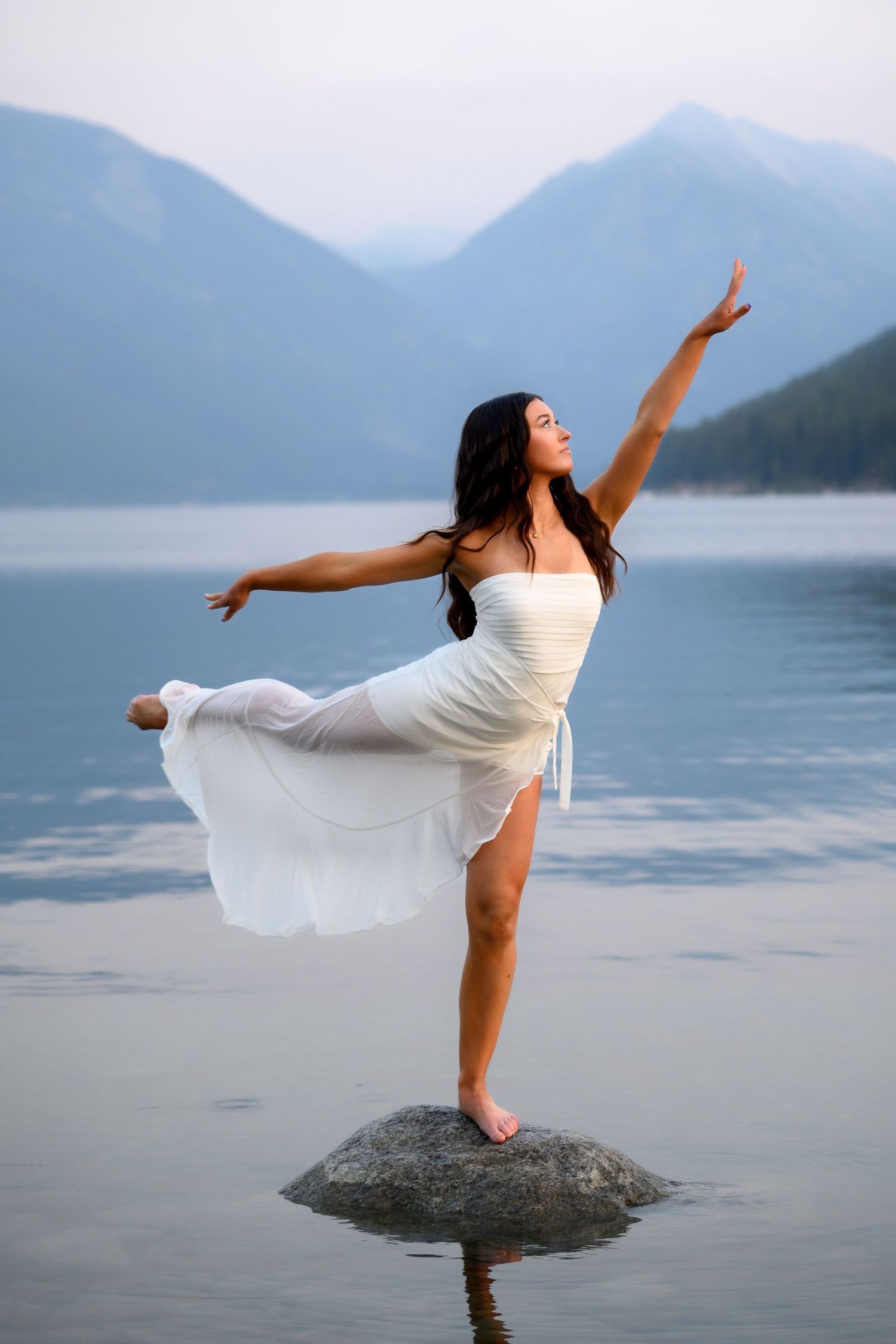 Dancer in flowing white dress strikes graceful pose on lake rock with misty mountains in background.
