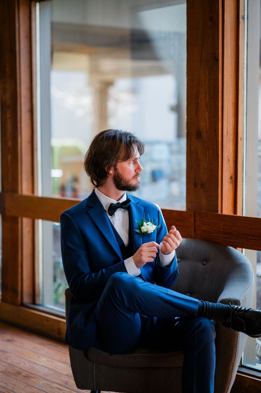A groom in a blue suit with boutonniere sits thoughtfully by large windows, adjusting his cufflinks before the wedding.