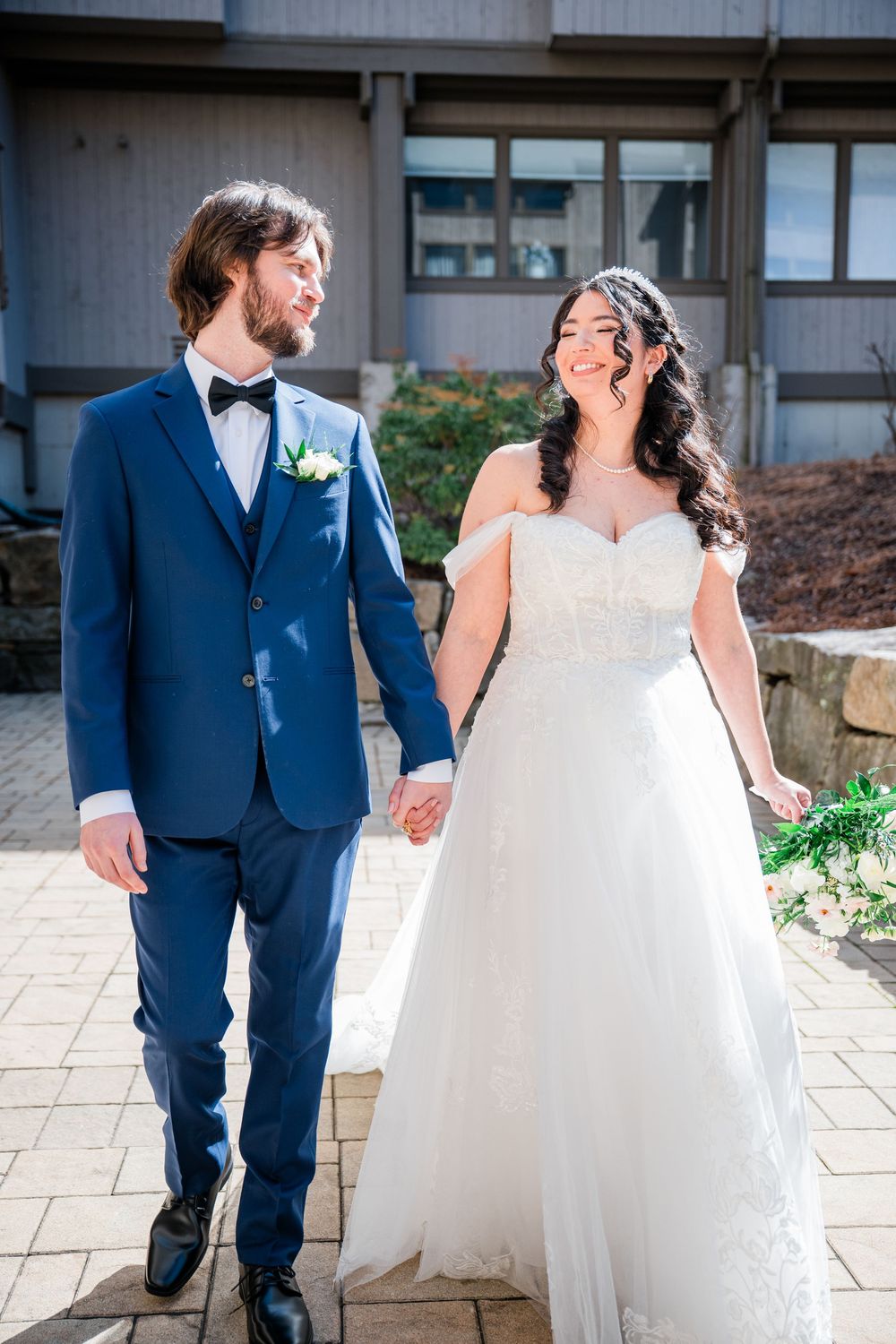 A smiling bride in a white off-shoulder gown holds hands with a groom in a navy blue tuxedo outdoors.