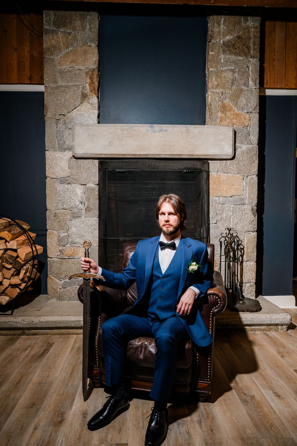 Man in blue suit with bow tie sits in leather chair holding a scepter before a stone fireplace.