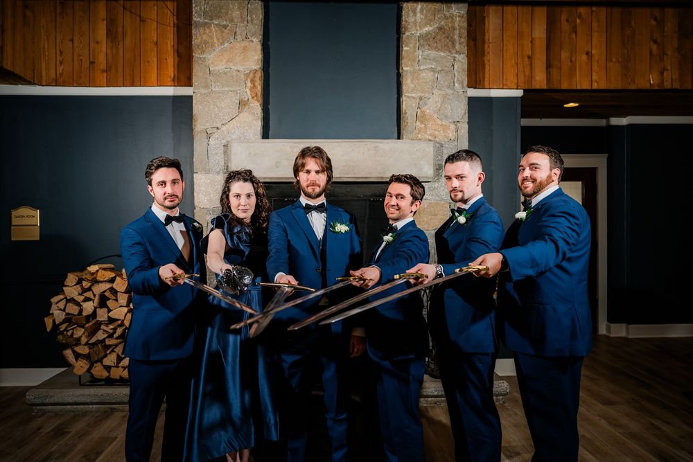 Wedding party in navy suits holding crossed swords indoors near stone fireplace.