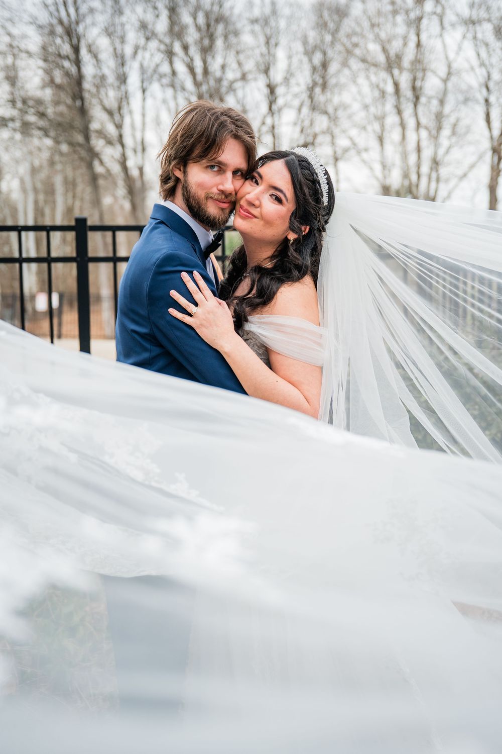 A bride and groom embrace outdoors, her flowing veil billowing around them, with bare winter trees in the background at the Heritage Hotel.