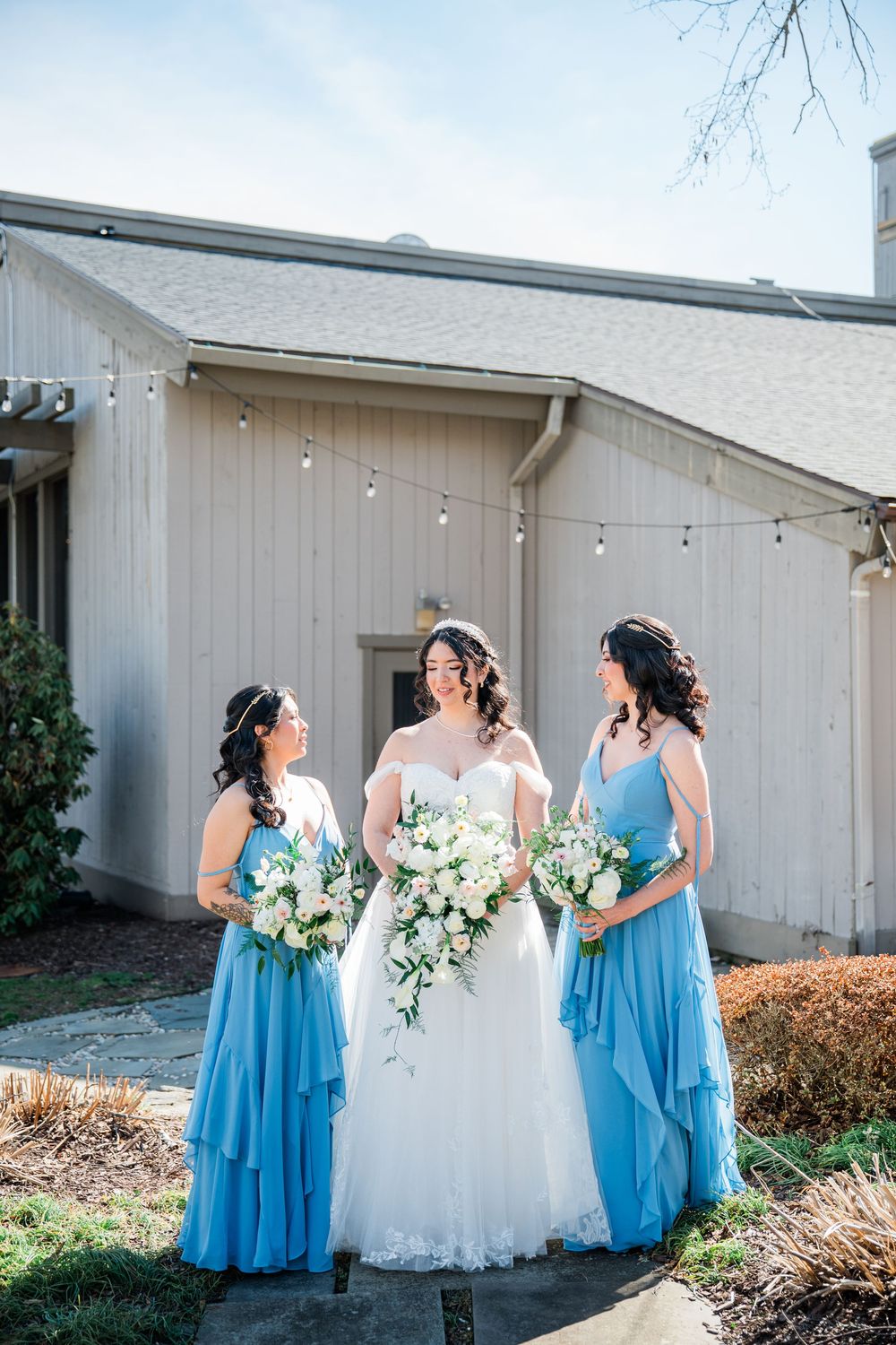 Bride in white gown stands with two bridesmaids in blue dresses holding white bouquets outside a barn venue.