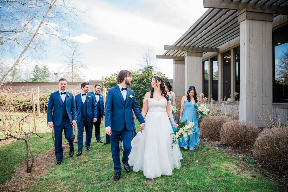 Wedding party walks outdoors, bride in white gown holding bouquet, groom in blue suit, groomsmen in navy, bridesmaid in blue dress.