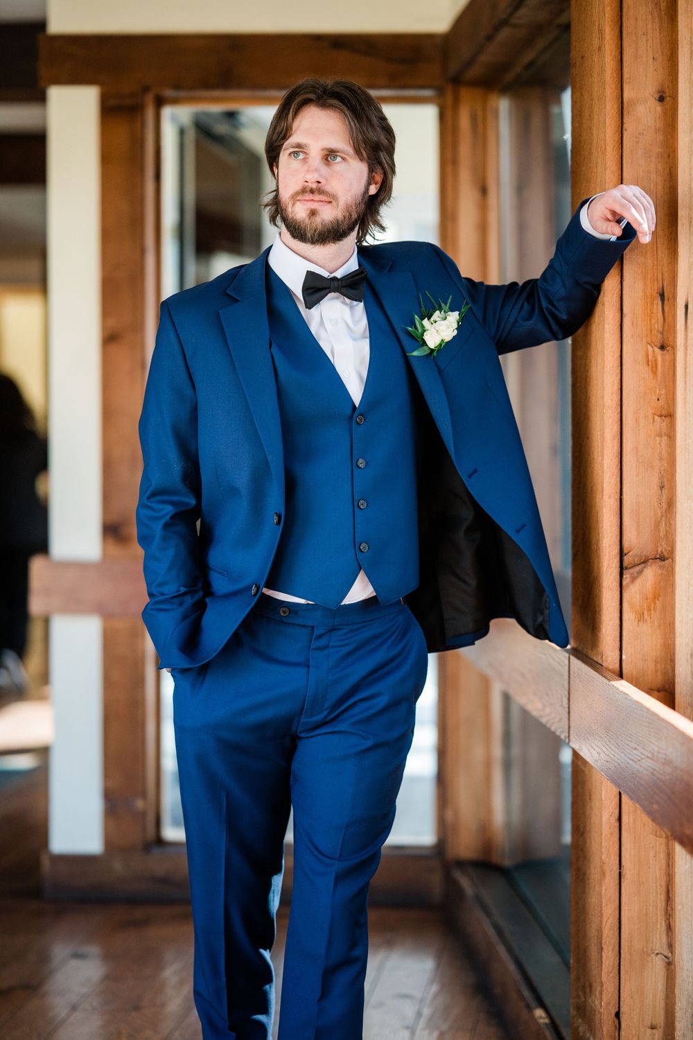 A dapper groom in a navy blue three-piece suit with bow tie and boutonniere leans against a wooden post.