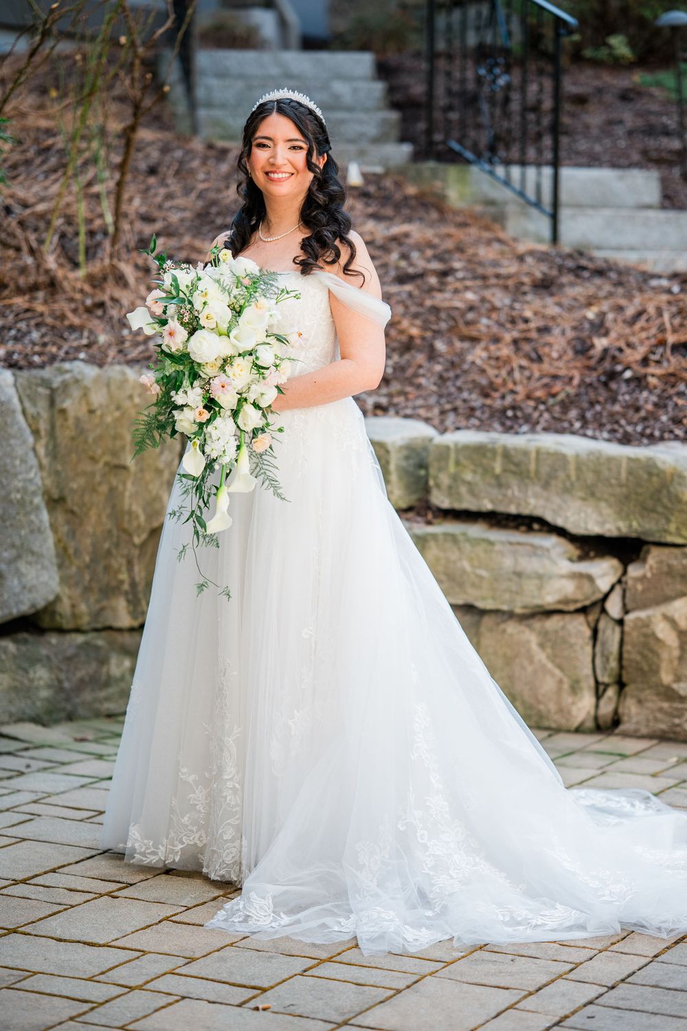 Bride in white lace off-shoulder gown holding cascading white floral bouquet, standing on stone patio outdoors.