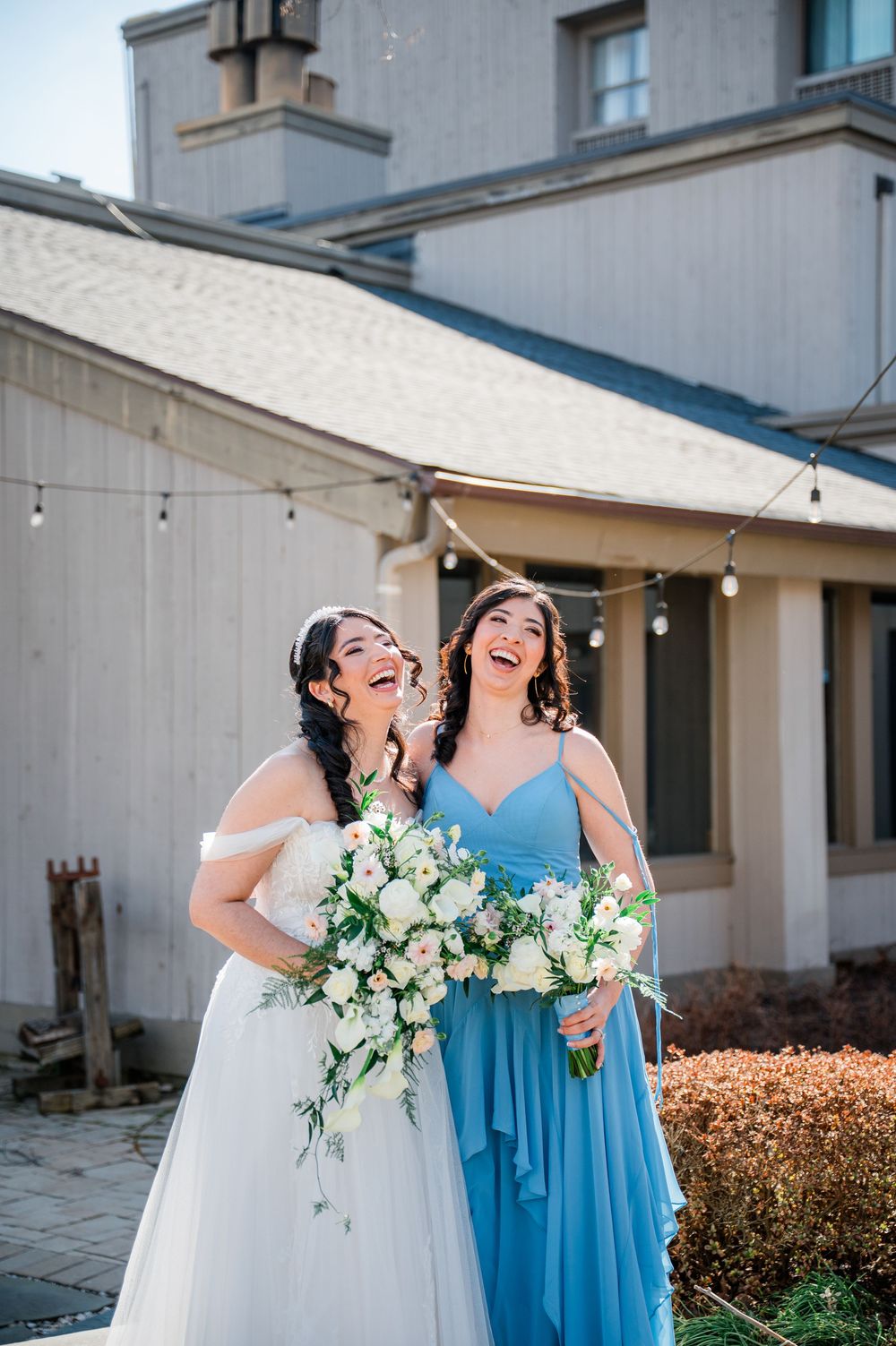 Bride in white gown and bridesmaid in blue dress laugh joyfully outdoors, holding white floral bouquets at wedding venue.