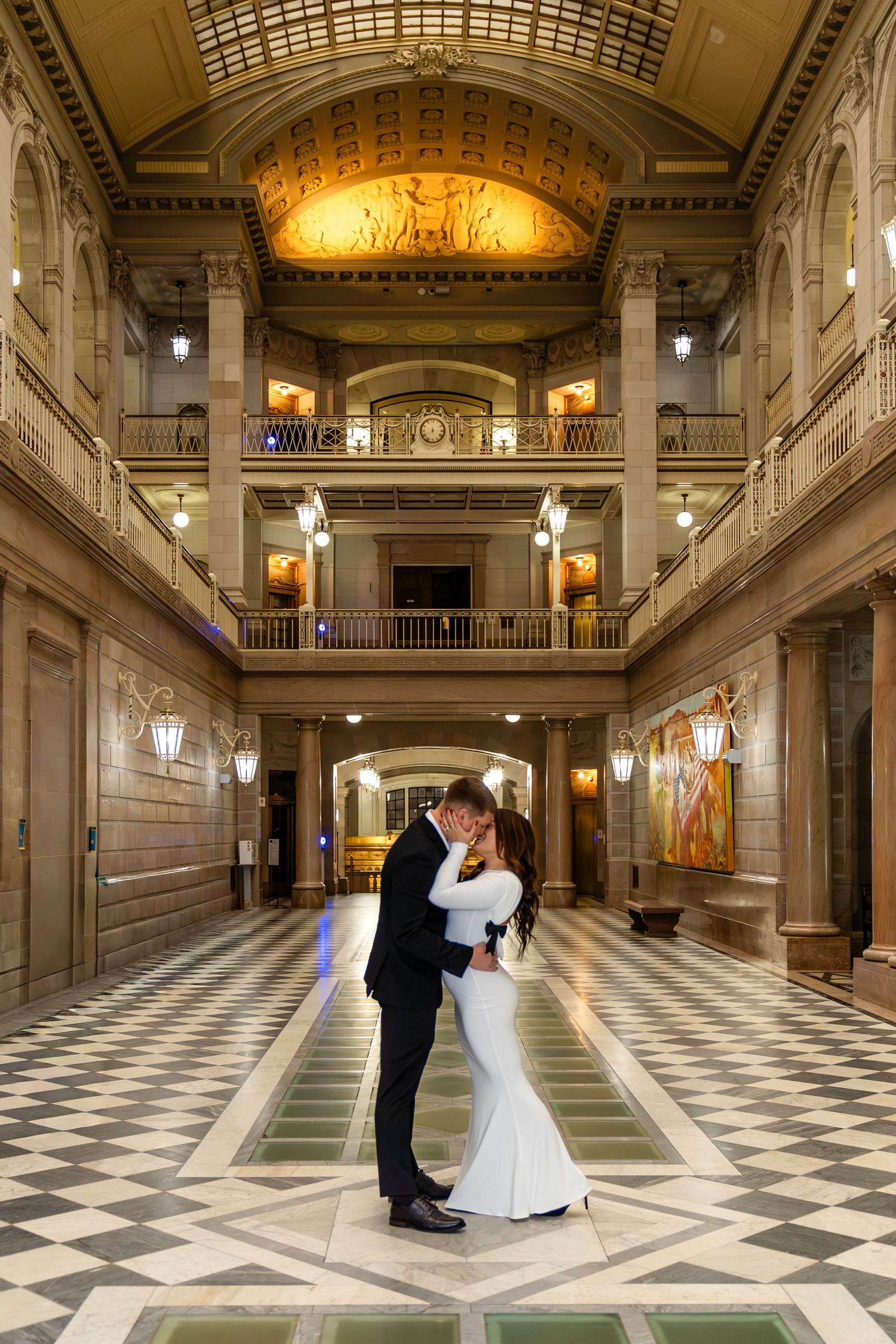 Bride and groom share romantic embrace in grand neoclassical building with ornate columns and checkered marble floor.