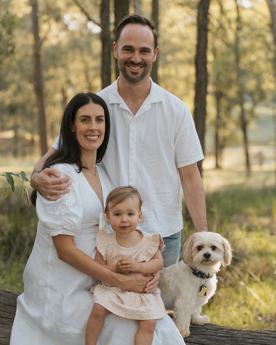 Family portrait of couple with toddler girl and small white dog outdoors in a sunlit forest in Rouse hill family photoshoot wit ha dog.