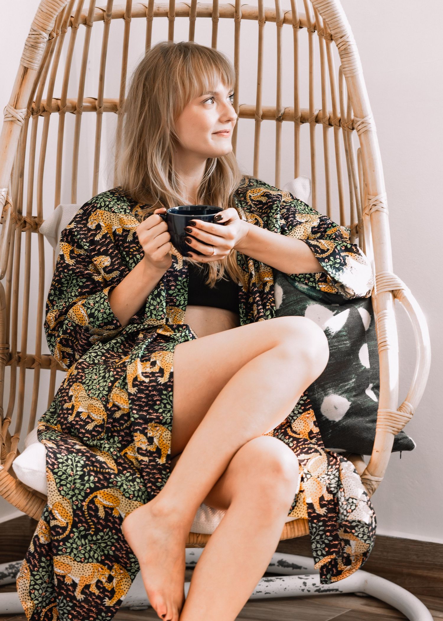 Woman in leopard print robe sitting in rattan egg chair holding a mug, relaxing in cozy home setting.