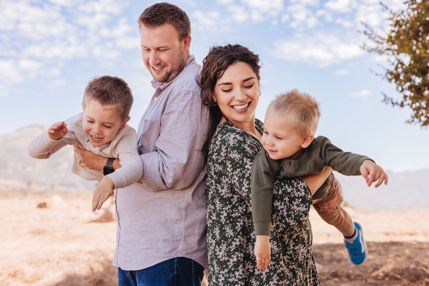 Family of four standing under a large oak tree in golden sunlight on a dry grassy hillside.