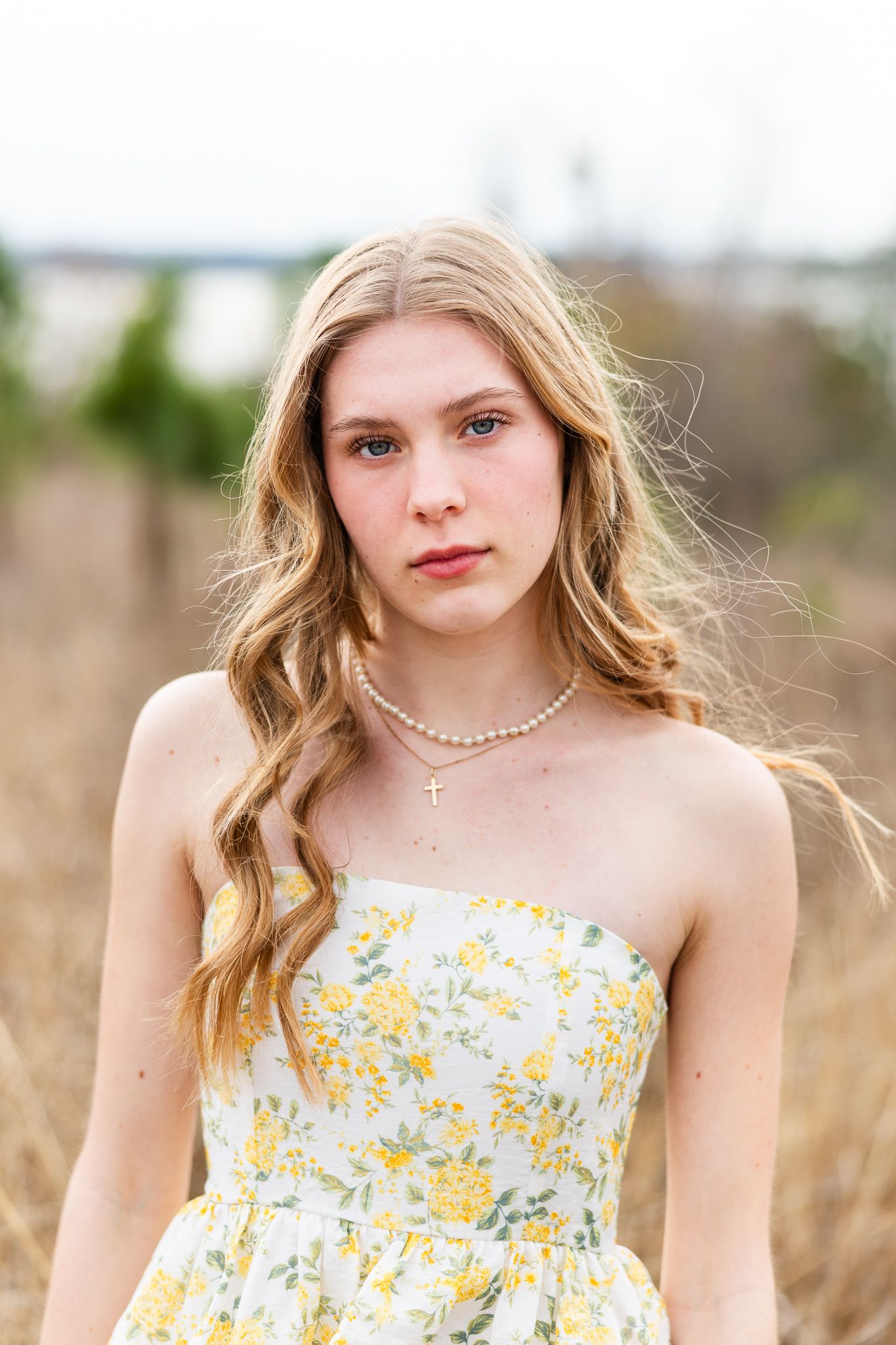 Young blonde woman in a floral tiered gown standing in dry grass field surrounded by pine trees.