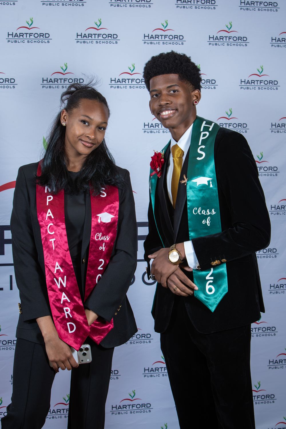 Two Hartford Public Schools graduates pose in black gowns with Class of 26 sashes at a graduation ceremony.