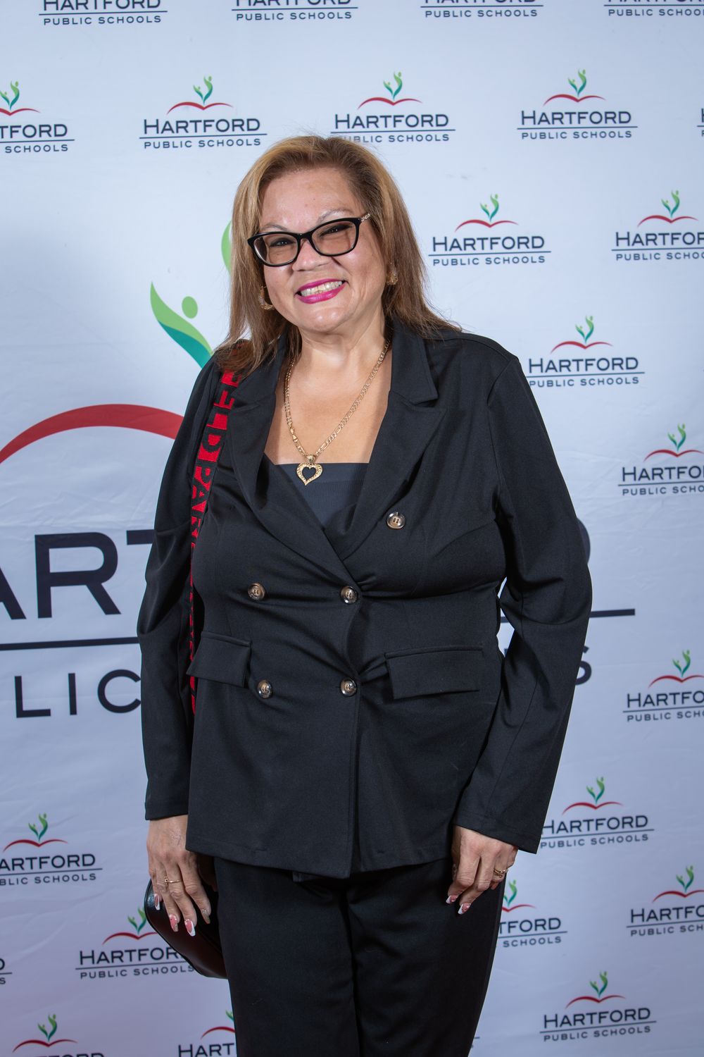 Woman in black blazer smiling at Hartford Public Schools event backdrop.