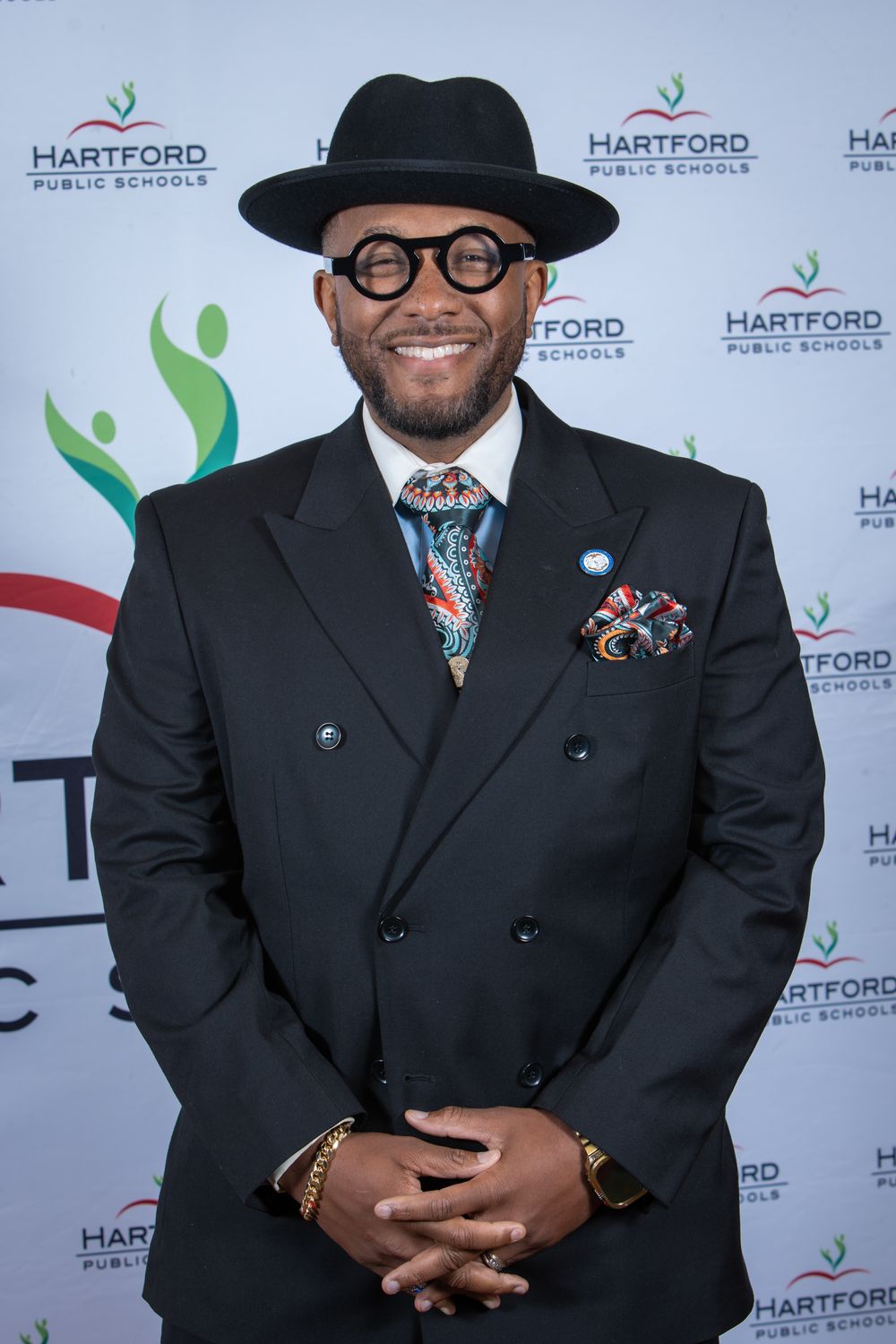 Man in black double-breasted suit, fedora, colorful tie and pocket square poses at Hartford Public Schools event.
