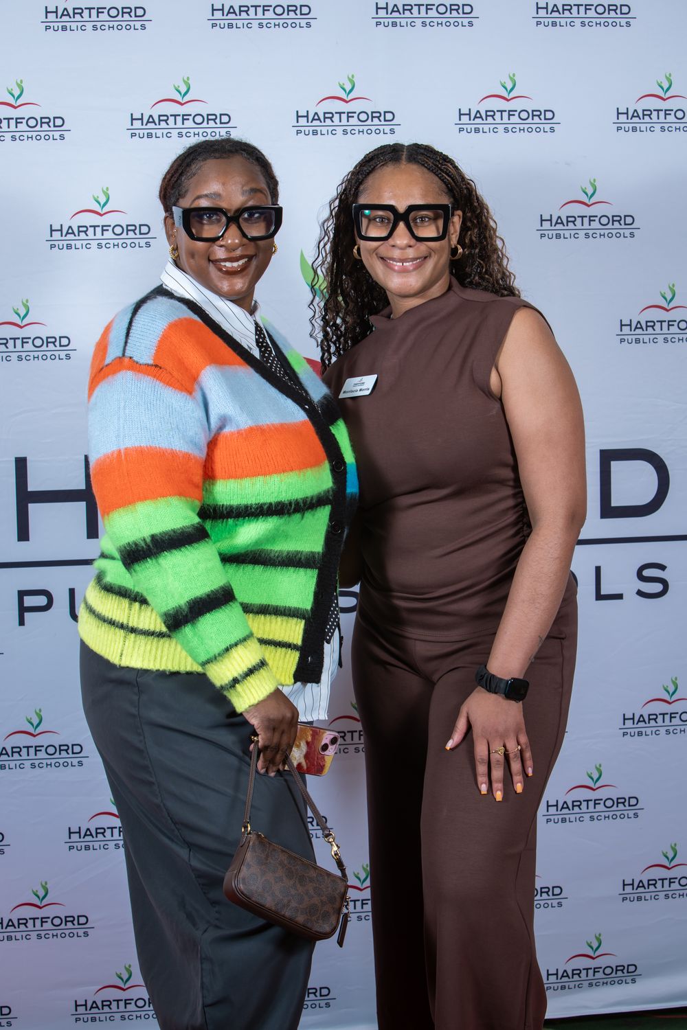 Two women in stylish outfits pose together at a Hartford Public Schools event backdrop.