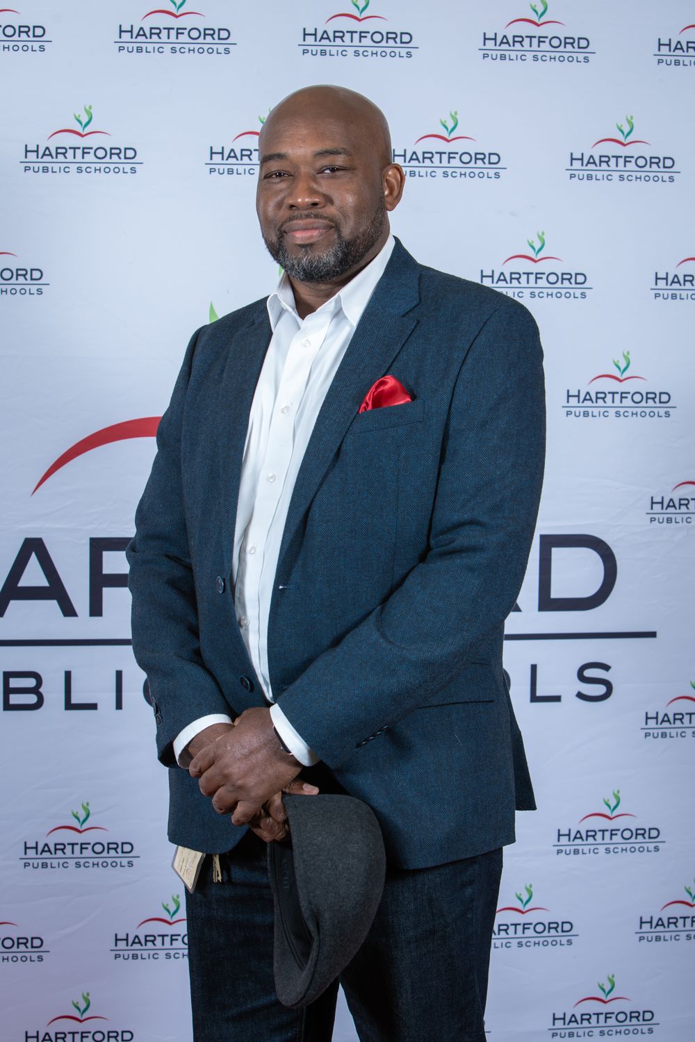 Man in navy blazer with red pocket square poses in front of Hartford Public Schools branded backdrop.