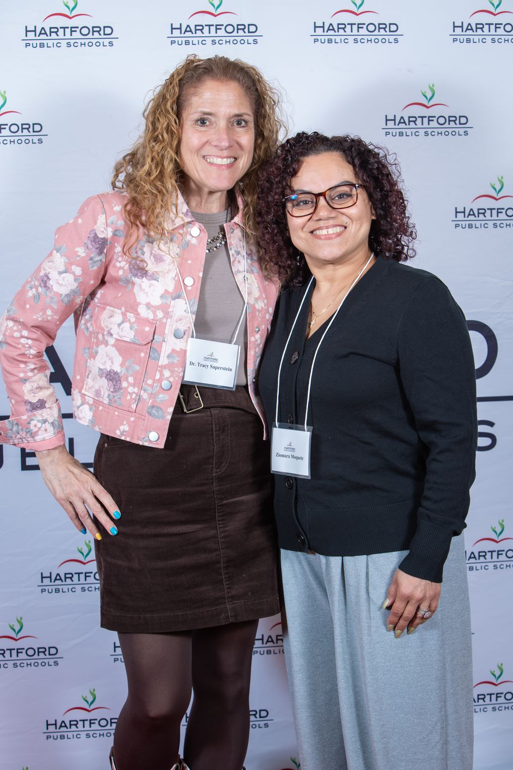 Two women smiling together in front of a Hartford Public Schools branded backdrop at a professional event.