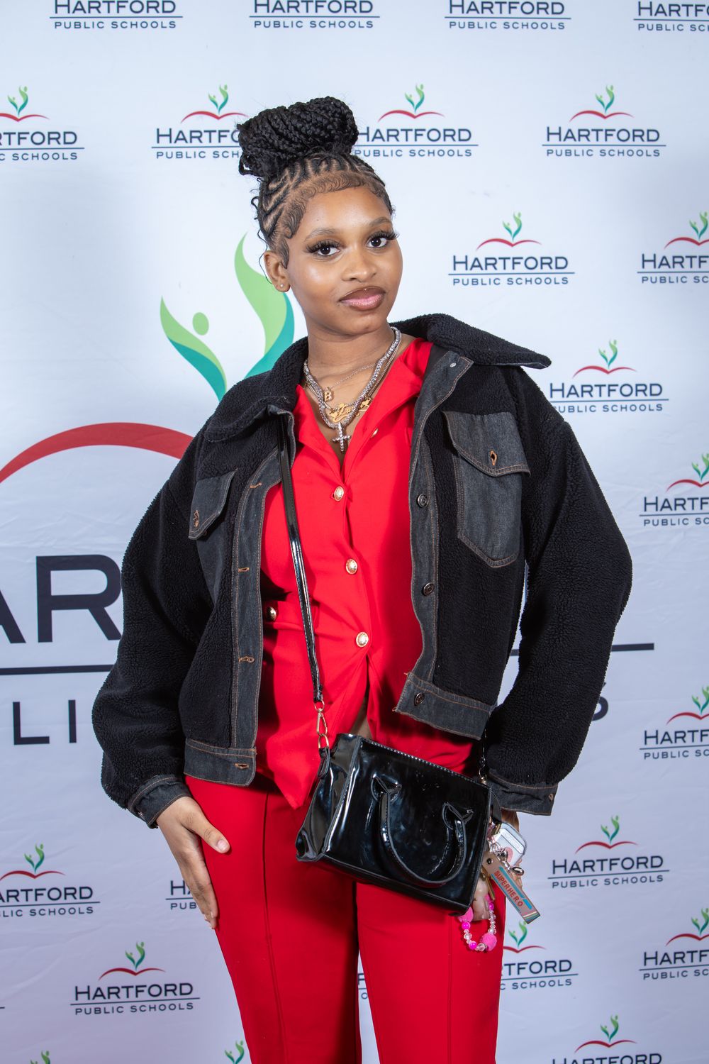 Young woman in red outfit and black denim jacket poses at Hartford Public Schools event backdrop.