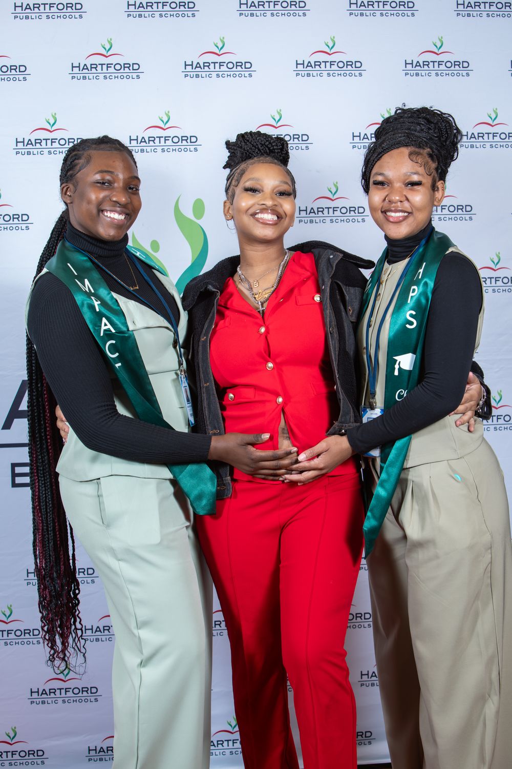 Three smiling students pose together at a Hartford Public Schools event backdrop, two wearing green sashes.