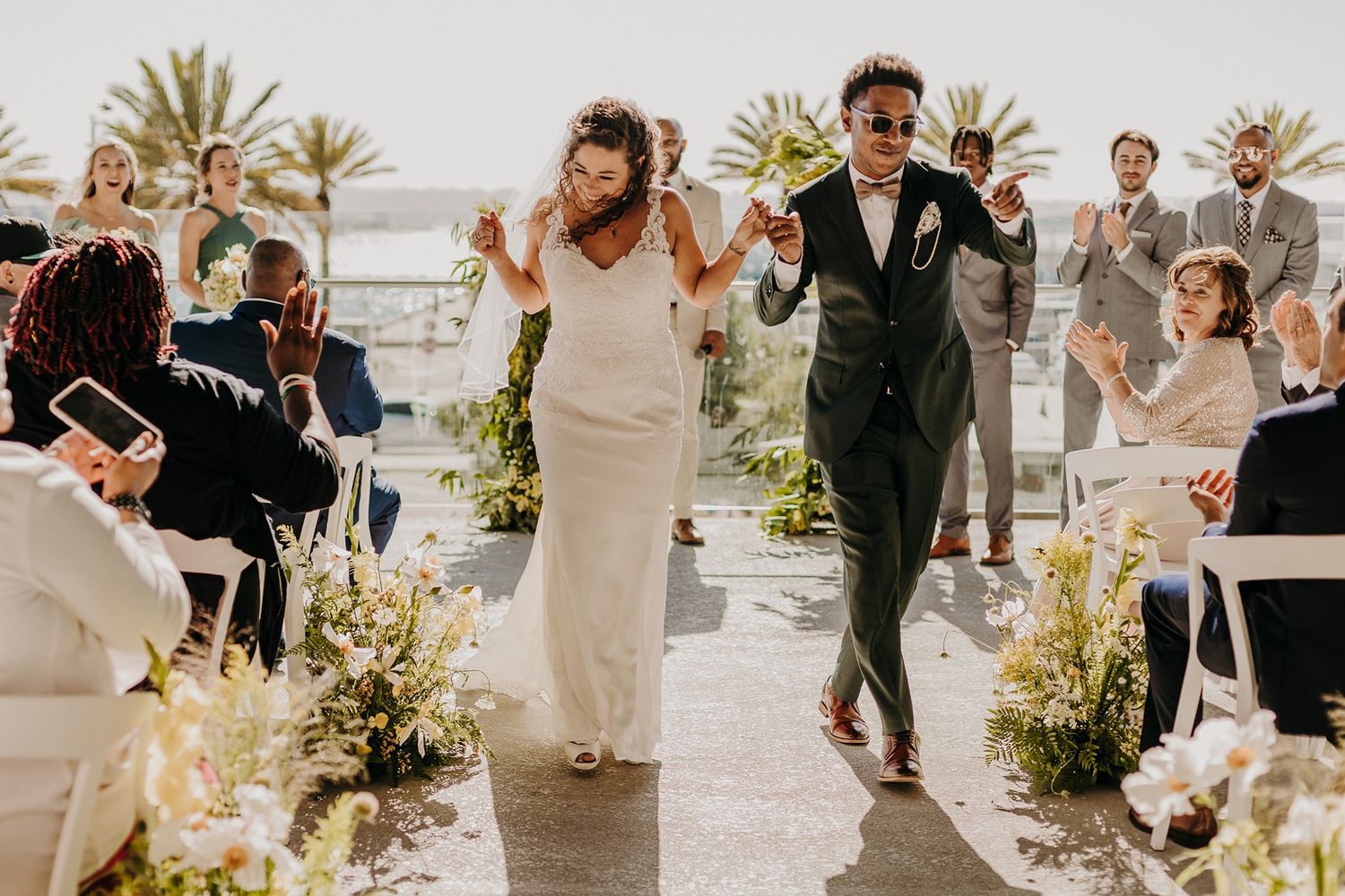 Joyful newlyweds walk down the aisle at an outdoor wedding ceremony, guests applauding as bride in white gown and groom celebrate.