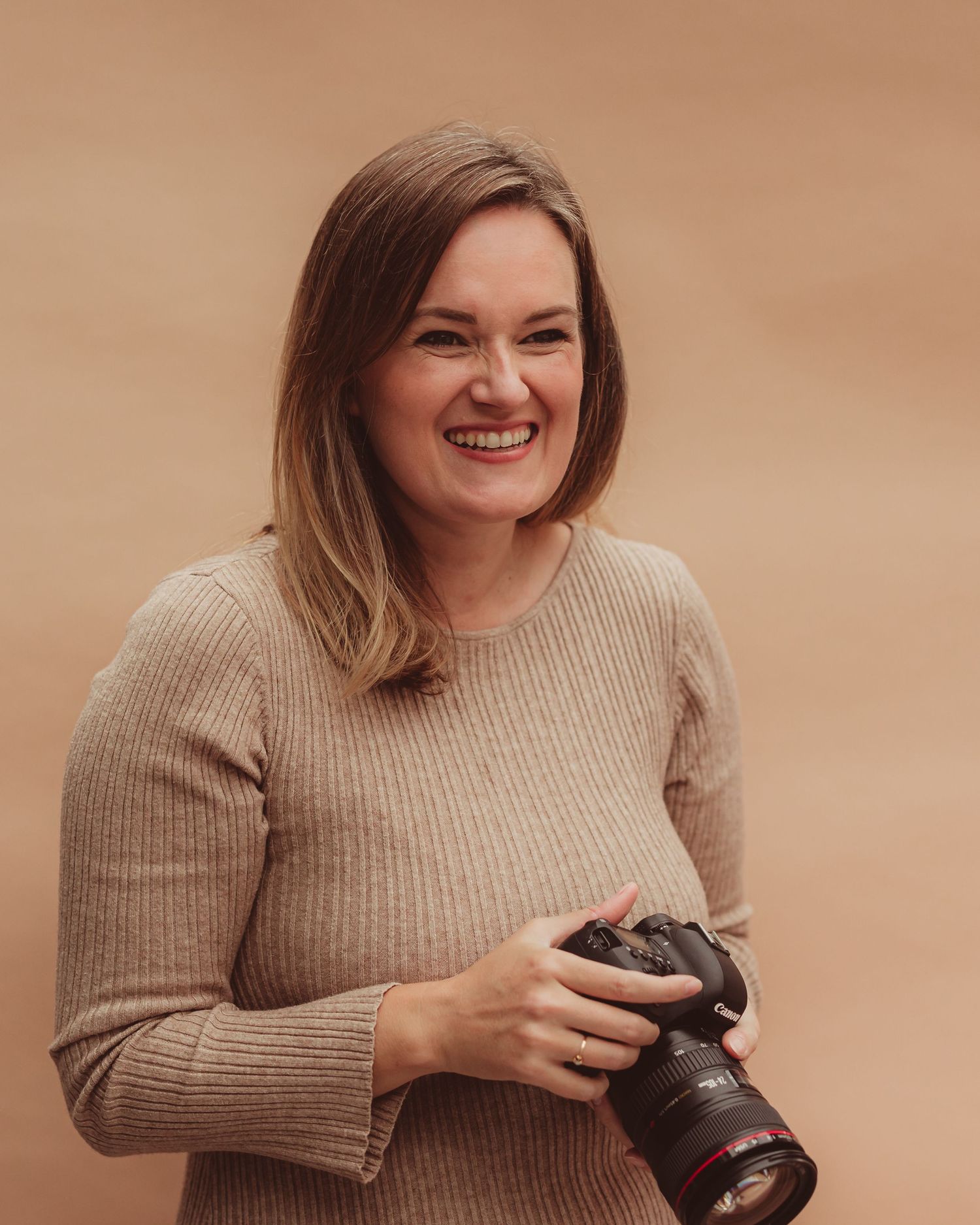 Smiling female photographer holding a Canon camera wearing a beige sweater against a warm brown background.