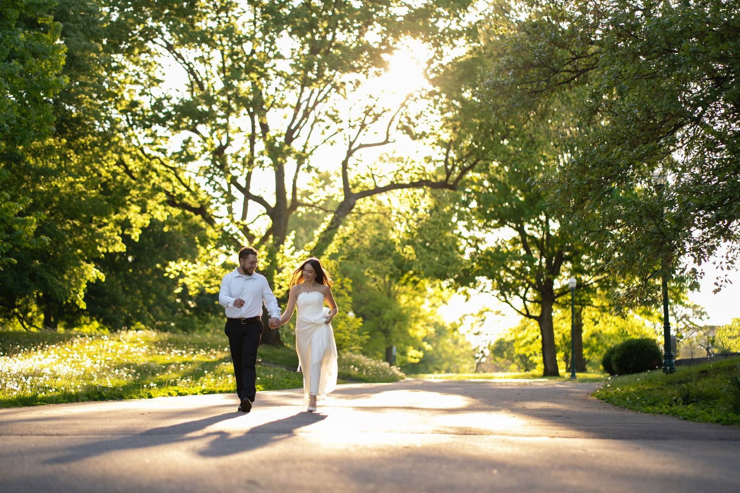 engaged Couple holding hands walking down sunlit tree-lined path in golden hour light at Franklin Park