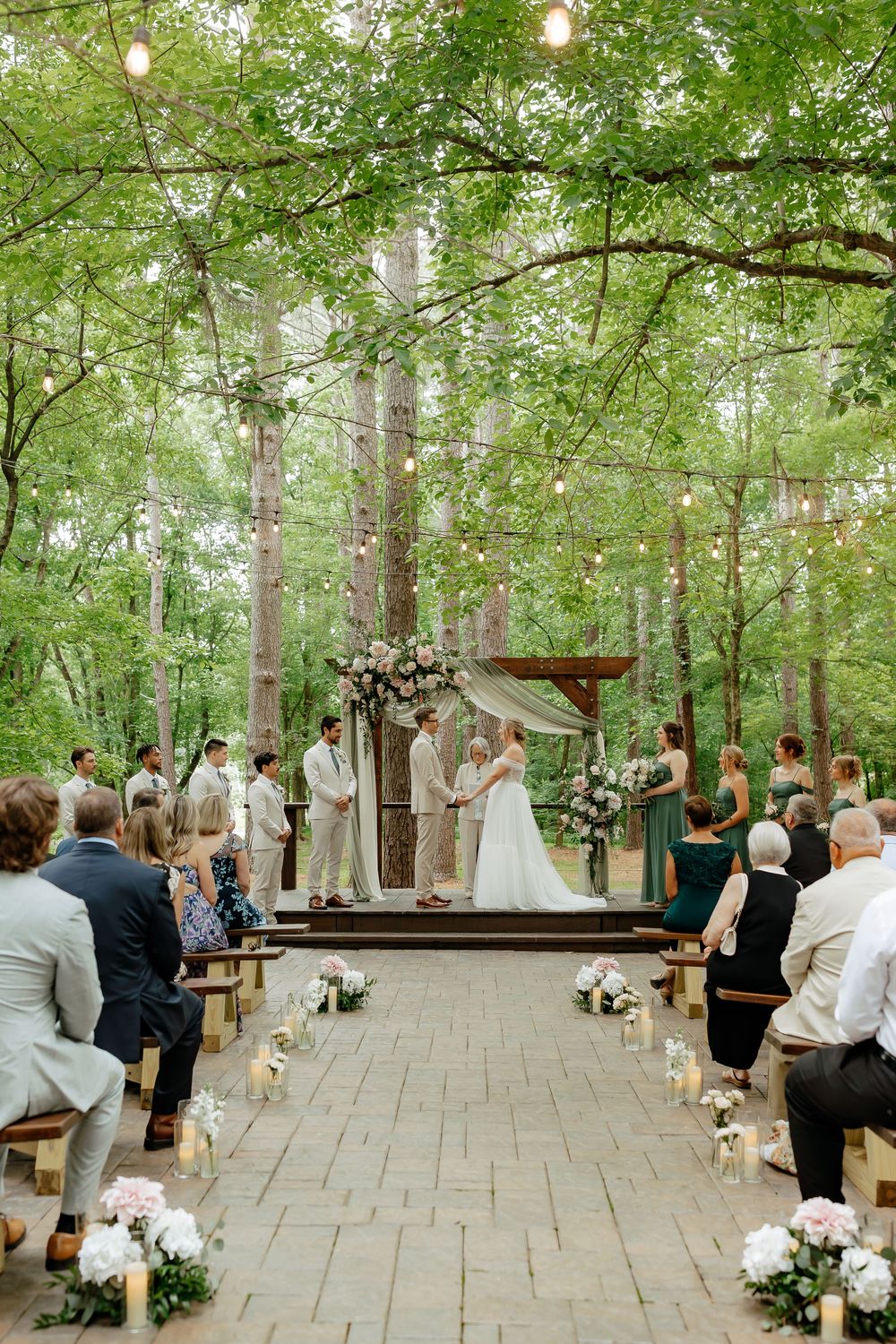Outdoor woodland wedding ceremony with string lights, floral arch, and guests seated on wooden benches among lush green trees.