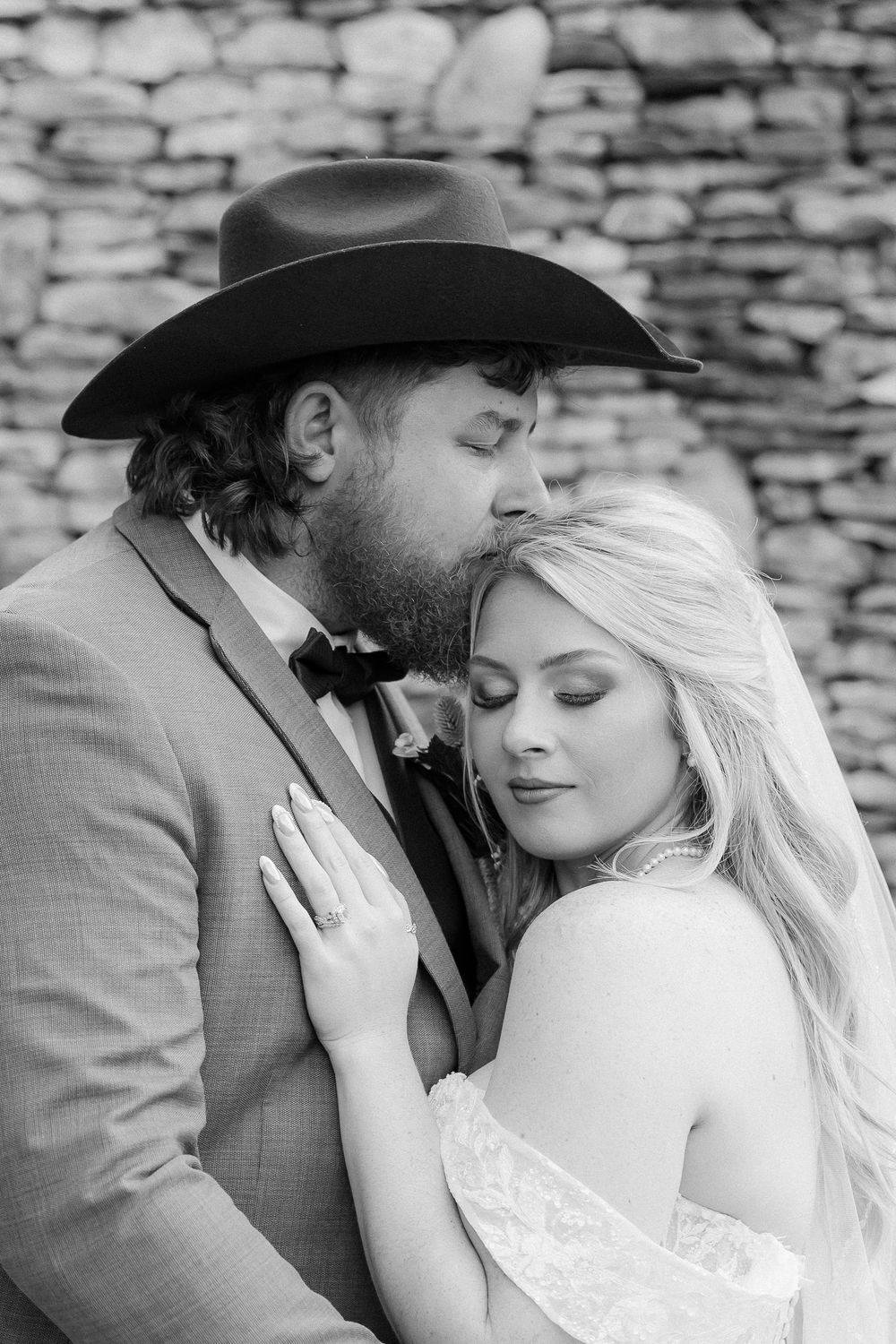 Black and white wedding photo of bearded groom in cowboy hat kissing bride's head as she smiles softly.