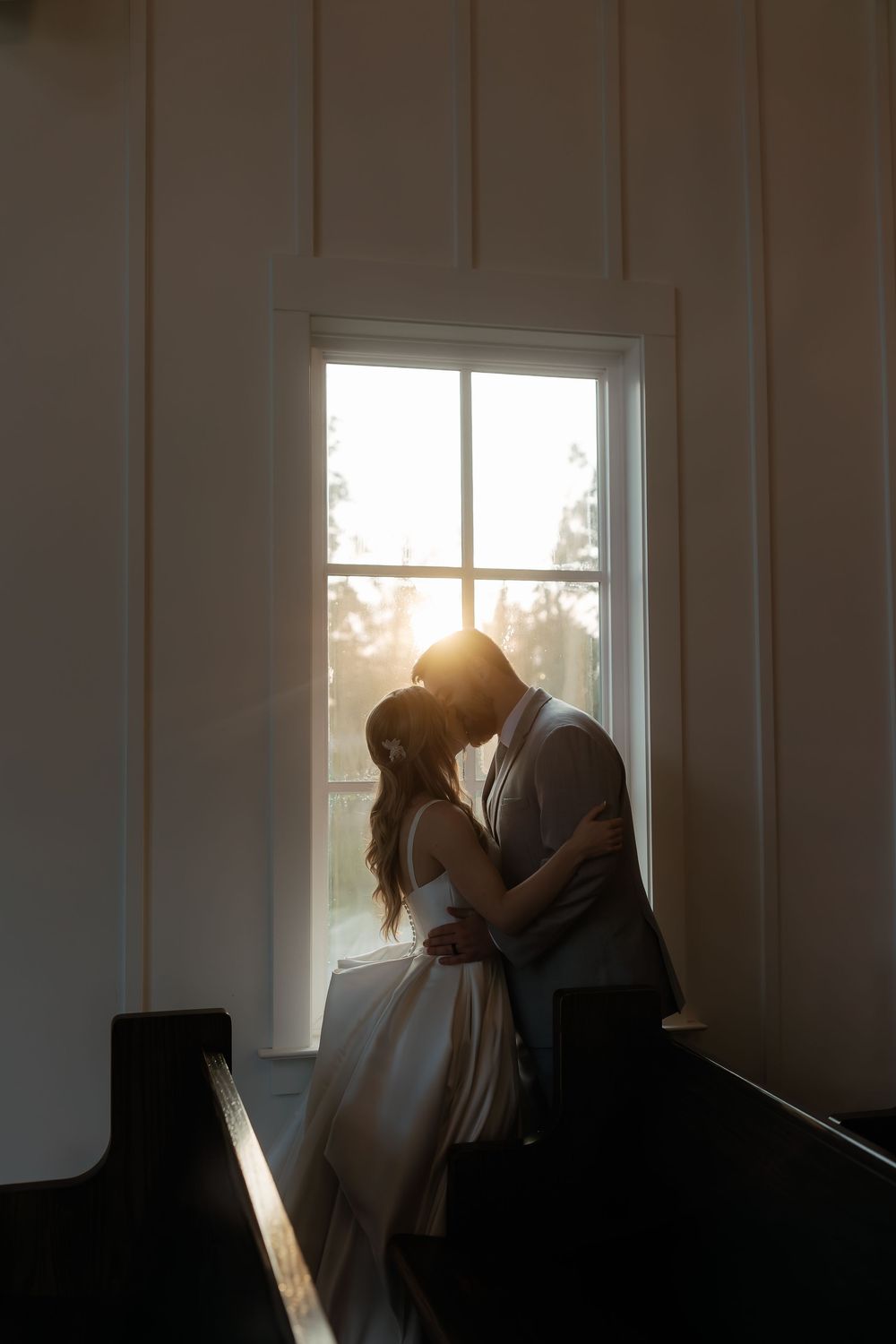 Bride and groom share a romantic moment backlit by golden sunset light through a church window.