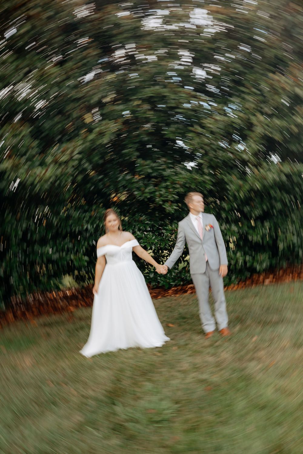 Bride in white off-shoulder gown and groom in gray suit hold hands outdoors with radial blur effect.