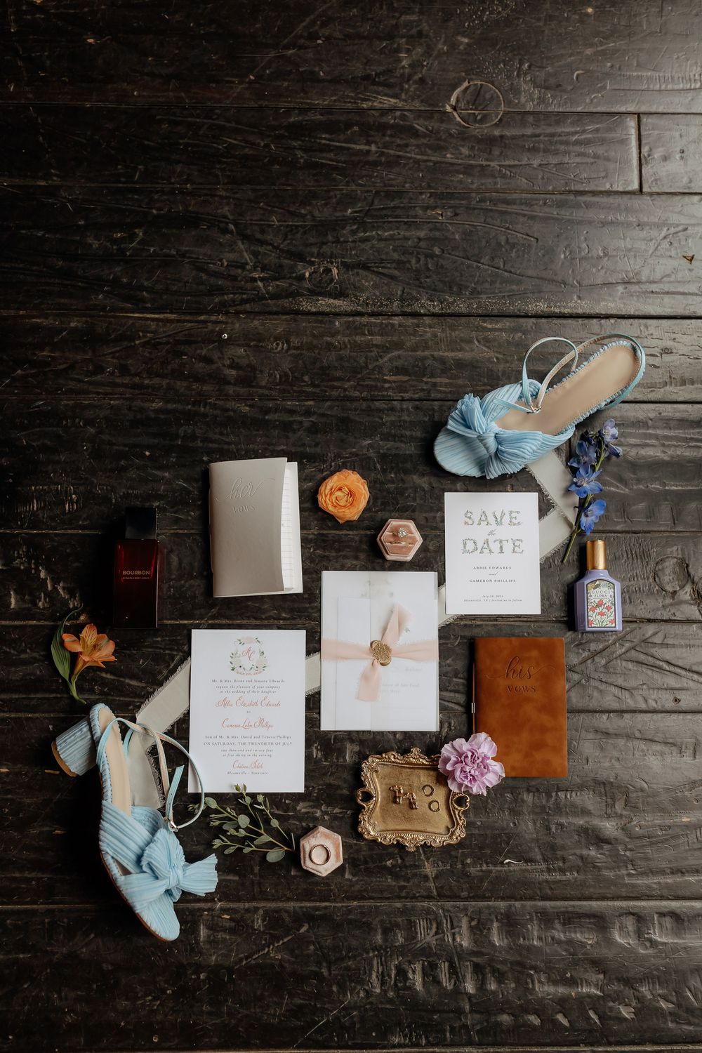 Elegant wedding flat lay with blue heels, save the date cards, floral accents, perfume, and accessories on dark wood.