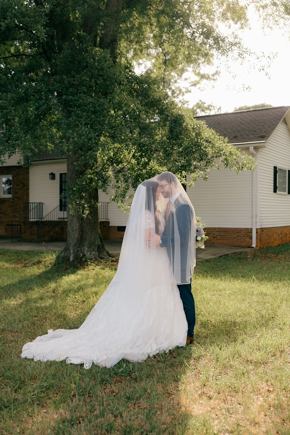 Bride and groom share a kiss under a flowing veil outdoors near a white house and large green tree.