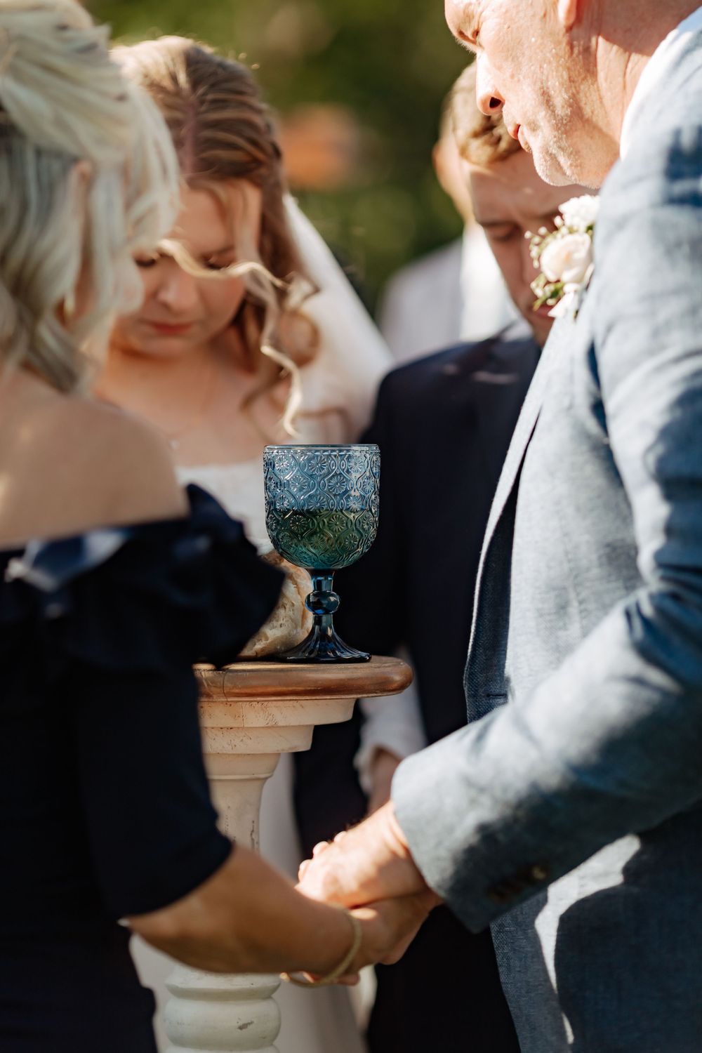 Bride and groom holding hands during outdoor wedding ceremony with decorative blue goblet on pedestal.
