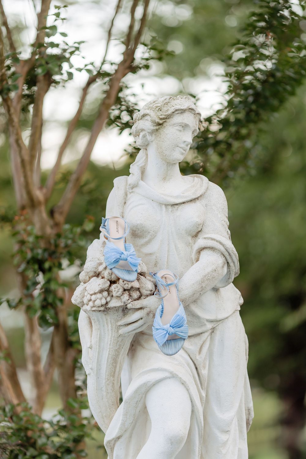White classical garden statue of a woman holding flowers, adorned with delicate blue baby shoes among greenery.