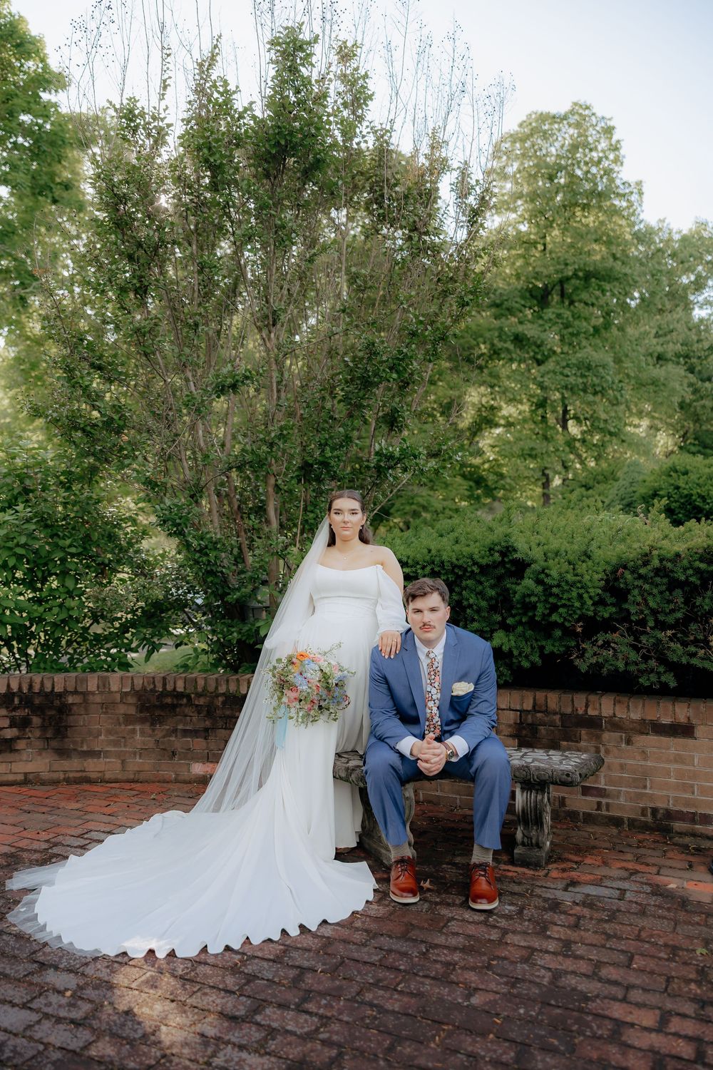 Bride in white gown with veil holds bouquet beside groom in blue suit seated on bench in lush garden.