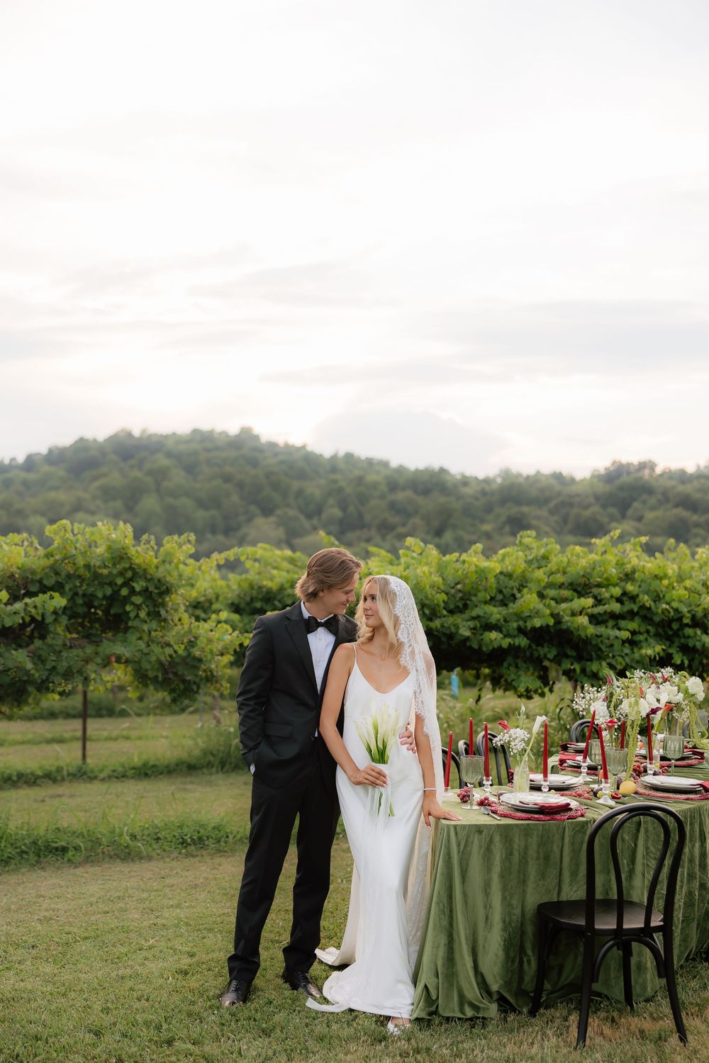 Bride and groom stand beside an elegant vineyard reception table with green velvet linen and red floral arrangements.