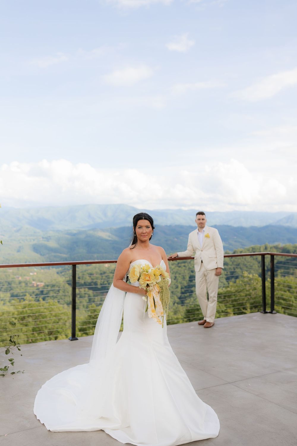 Bride in white gown holds yellow bouquet on mountain overlook, groom in tan suit stands behind with scenic views.