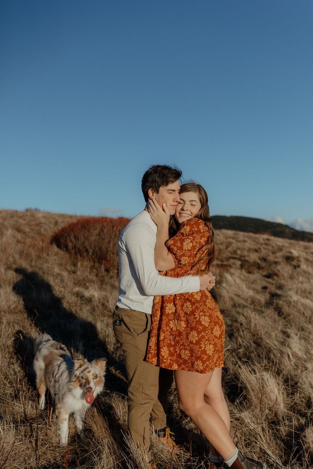 Couple embracing in golden field at sunset, woman in rust floral dress smiling, man kissing her cheek, dog nearby.