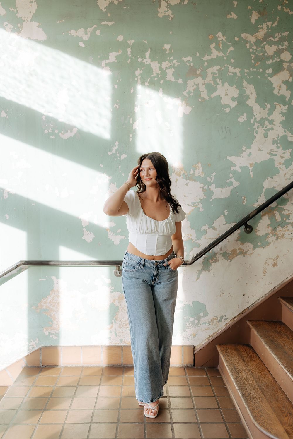 Young woman in white crop top and wide-leg jeans poses on stairs near peeling mint green wall in natural light.