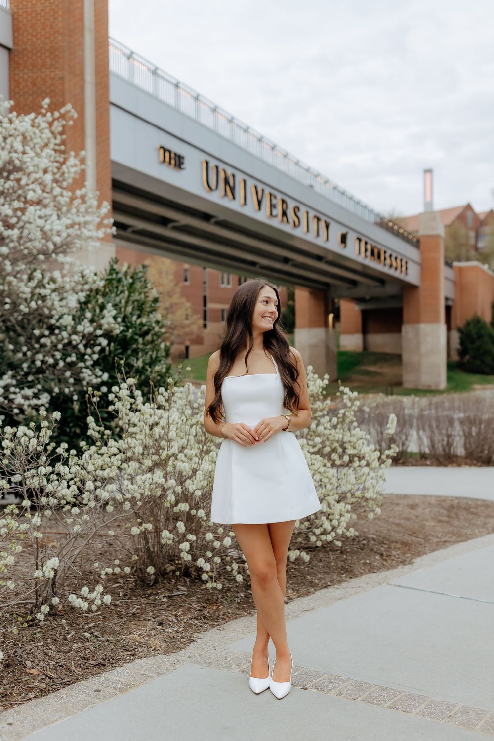 Young woman in white dress and heels poses in front of University of Tennessee sign amid spring blooms.