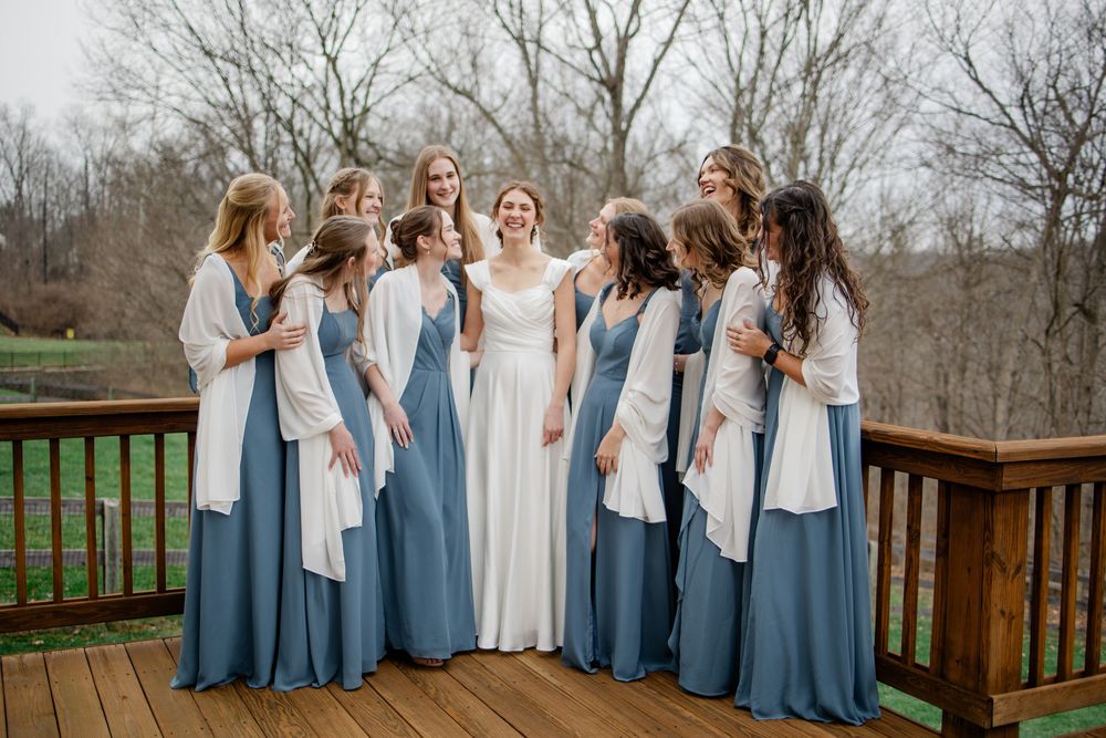 Bride in white gown surrounded by bridesmaids in dusty blue dresses on a wooden deck outdoors.