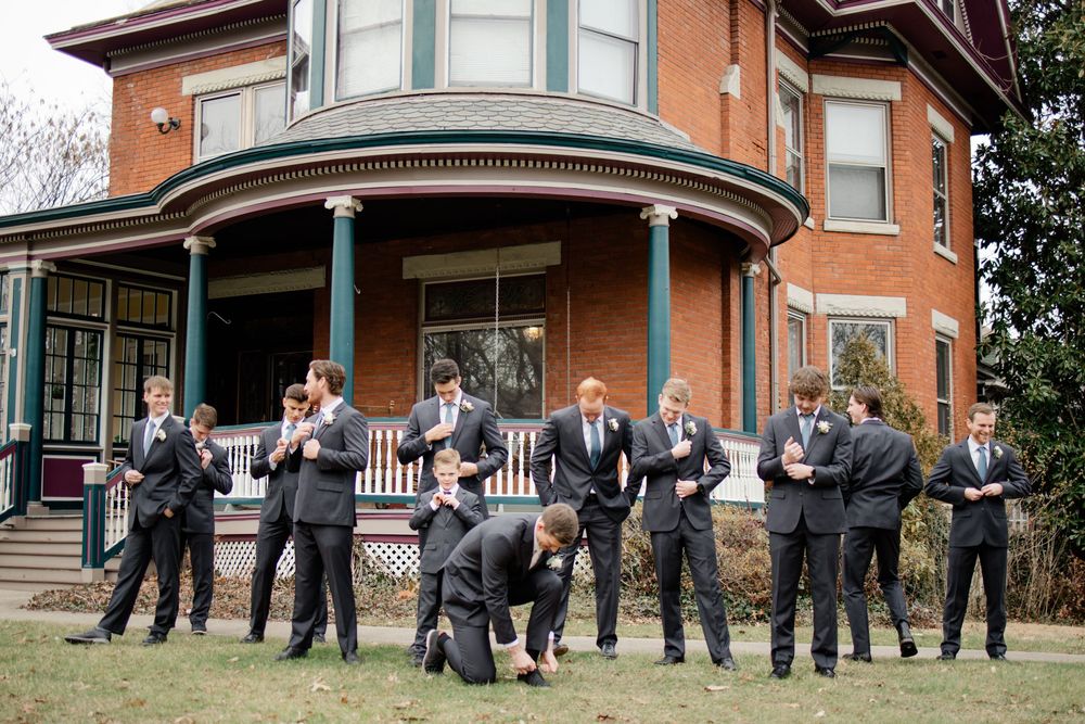 Groomsmen in dark suits pose playfully outside a brick Victorian house with a wraparound porch at a wedding.