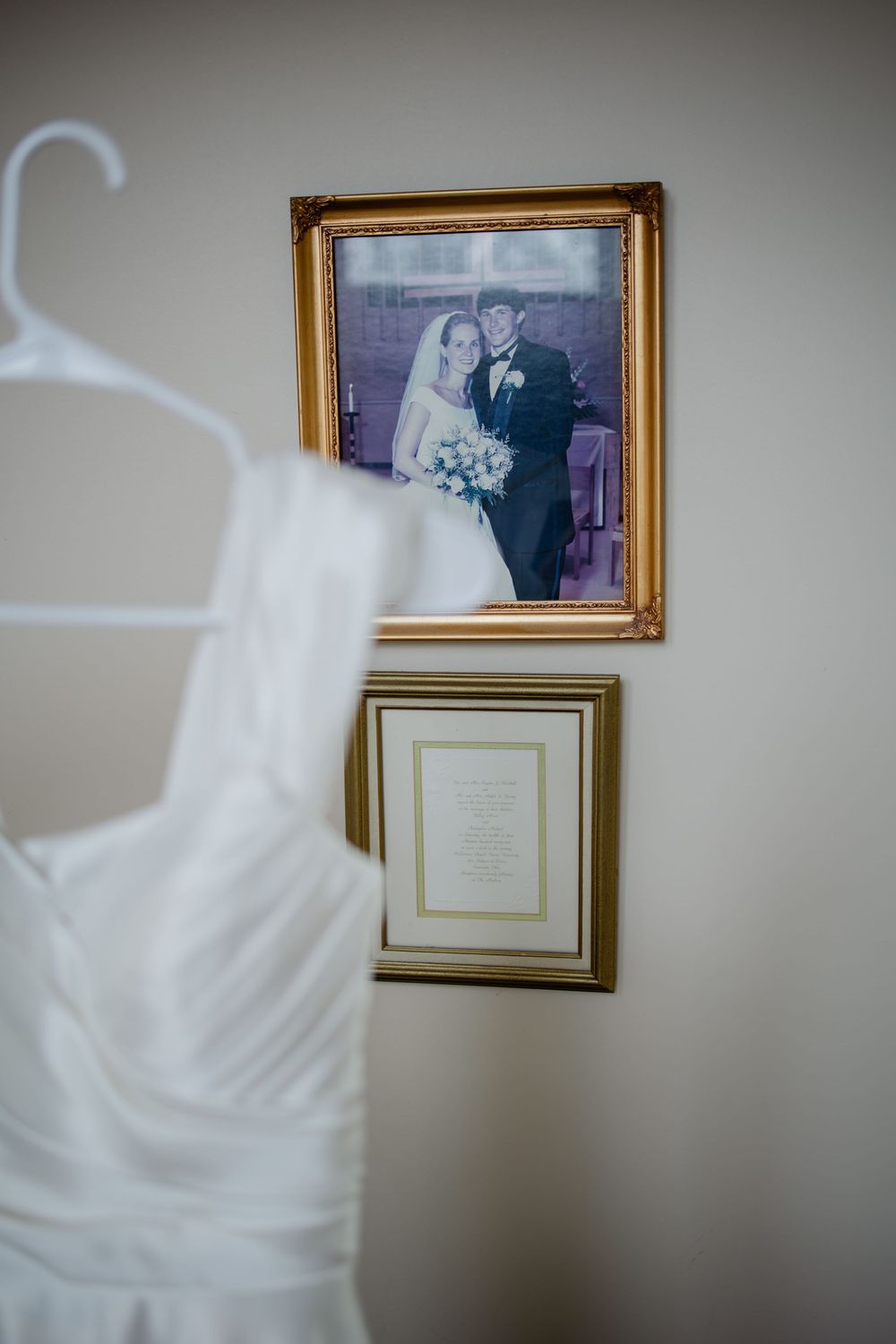 White wedding dress hanging near framed wedding photo of bride and groom on wall with gold frames.